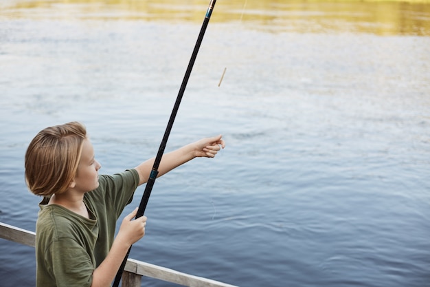 Attraktiver kleiner Kerl, der Angelrute zum Fluss wirft, große Fische fangen will, Wochenende in der Natur verbringen, in der Nähe von Fluss oder See, sehr konzentriert.