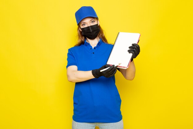 Attraktiver Kurier der weiblichen Vorderansicht in der blauen Kappe des blauen Poloshirts und der Jeans in der schwarzen Schutzmaske der schwarzen Handschuhe, die Stift und Notizbuch auf dem gelben Hintergrundlebensmittelservicejob hält