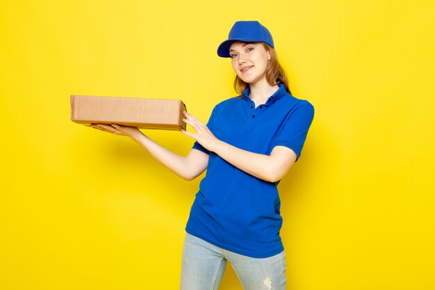 Attraktiver Kurier der weiblichen Vorderansicht in der blauen Kappe des blauen Poloshirts und im lächelnden Haltepaket der Jeans auf dem gelben Hintergrundlebensmittelservicejob