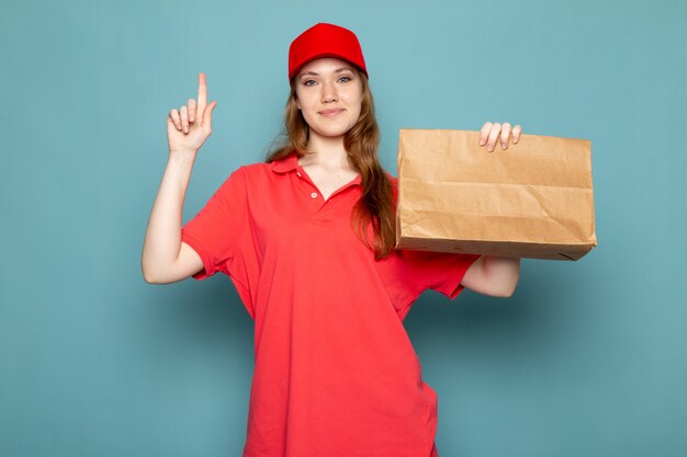 Attraktiver Kurier der weiblichen Vorderansicht in der roten Kappe des roten Poloshirts, die braunes Paket hält, das auf dem blauen Hintergrundlebensmittelservicejob lächelt