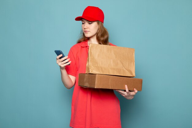 Attraktiver Kurier der weiblichen Vorderansicht in der roten Kappe des roten Poloshirts, die braunes Paket unter Verwendung des Telefons auf dem blauen Hintergrundlebensmittelservicejob hält