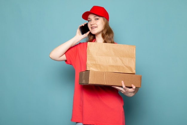 Attraktiver Kurier der weiblichen Vorderansicht in der roten Kappe des roten Poloshirts, die das lächelnde braune Paket hält, das am Telefon auf dem blauen Hintergrundlebensmittelservicejob spricht