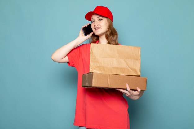 Attraktiver Kurier der weiblichen Vorderansicht in der roten Kappe des roten Poloshirts, die das lächelnde braune Paket hält, das am Telefon auf dem blauen Hintergrundlebensmittelservicejob spricht