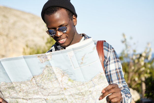 Attraktiver lächelnder junger afroamerikanischer Hipster im stilvollen schwarzen Hut und in der Sonnenbrille, die große Papierkarte konsultieren, während Sie im fremden Land besichtigen, nach der richtigen Richtung suchen, Sommerferien genießen