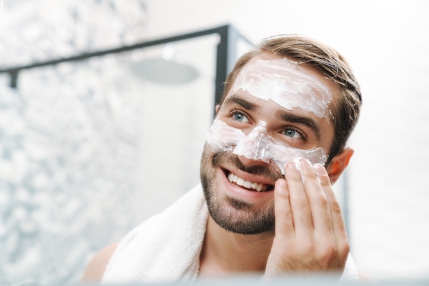 Attraktiver lächelnder junger mann ohne hemd, der gesichtscreme aufträgt, während er am badezimmerspiegel steht