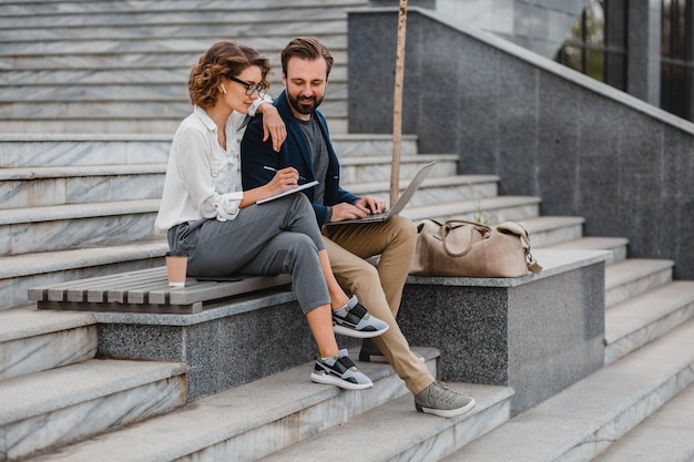 Attraktiver lächelnder Mann und Frau, die auf Treppen im städtischen Stadtzentrum sitzen und sich Notizen machen