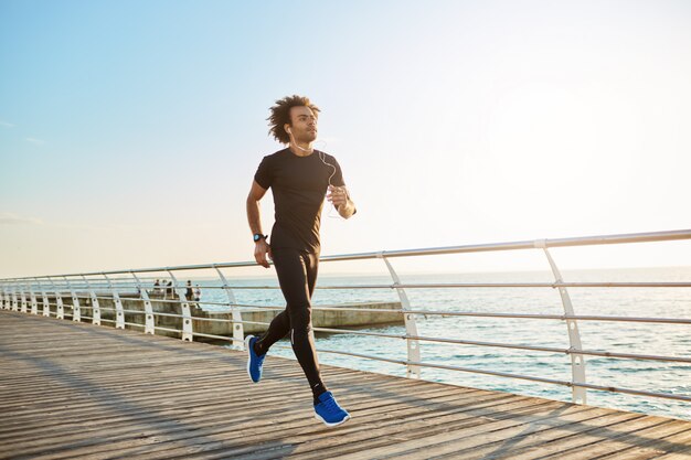 Attraktiver männlicher Athlet, der stilvolle schwarze Sportkleidung und blaue Turnschuhe trägt. Figur des Mannathleten, der Cardio-Laufübungen am sonnigen Sommermorgen tut.