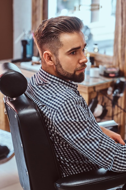 Attraktiver nachdenklicher Mann wartet darauf, dass er an der Reihe ist, um im geschäftigen Friseursalon einen Haarschnitt zu bekommen.