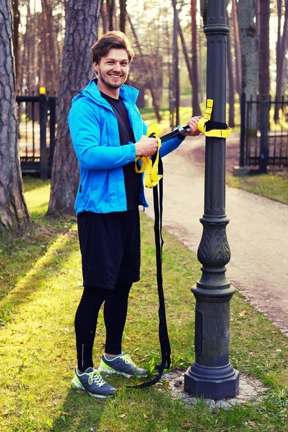 Attraktiver positiver Mann in einem blauen Regenmantel, der in einem Park mit TRX-Fitnessstreifen trainiert.