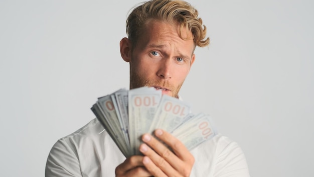 Attraktiver, reicher, blonder, bärtiger Geschäftsmann, der zuversichtlich aussieht und Geldbündel vor der Kamera vor weißem Hintergrund zeigt