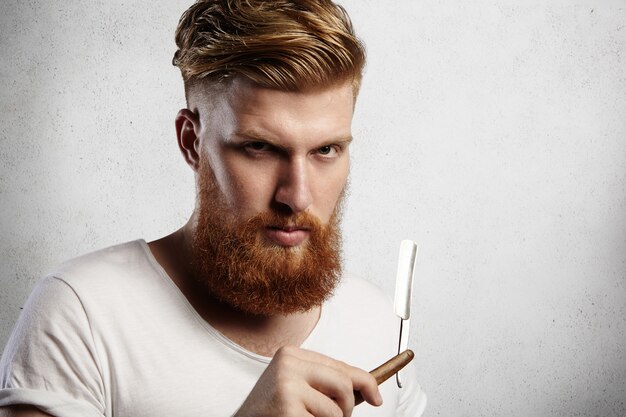Attraktiver Rotschopf-Hipster-Friseur mit stilvollem Haarschnitt und dickem Bart, der ein Rasiermesser mit ernstem Gesichtsausdruck hält.