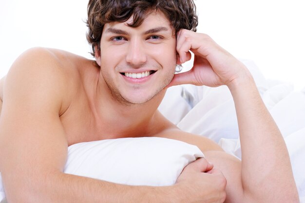 Attraktiver sexy lächelnder junger nackter Mann, der im Bett mit Kissen liegt