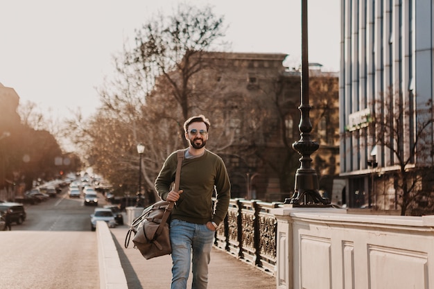 Attraktiver stilvoller Hipster-Mann, der in der Stadtstraße mit Ledertasche geht, die Sweatshot und Sonnenbrille trägt, Stadtstil-Trend, sonniger Tag