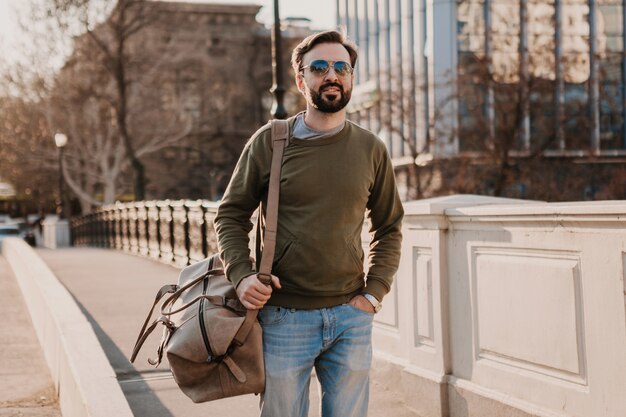 Attraktiver stilvoller Hipster-Mann, der in der Stadtstraße mit Ledertasche geht, die Sweatshot und Sonnenbrille trägt, Stadtstil-Trend, sonniger Tag