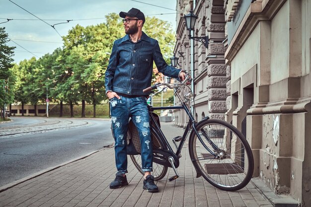Attraktiver, stylischer Hipster posiert neben seinem Fahrrad in der Nähe des Gebäudes. Er hält sein Fahrrad. Der Mann trägt Denim, Mütze und Brille. Eine Hand steckte er in seine Tasche.