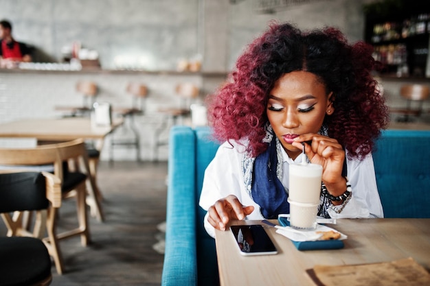 Attraktives afrikanisch-amerikanisches lockiges Mädchen, das im Café sitzt und Latte trinkt und auf das Handy schaut