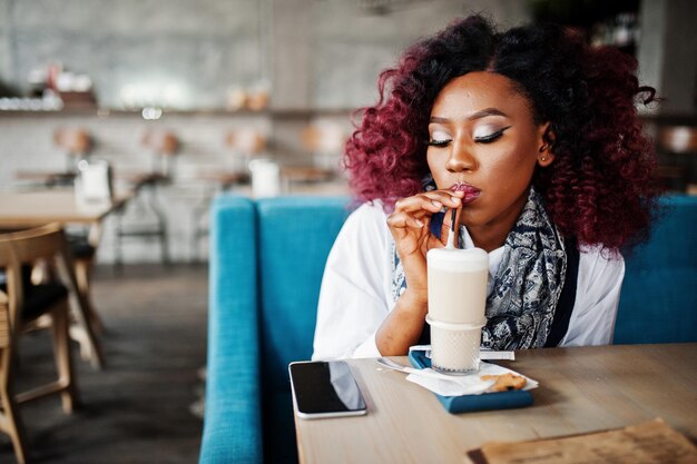 Attraktives afroamerikanisches lockiges Mädchen, das im Café sitzt und Latte trinkt