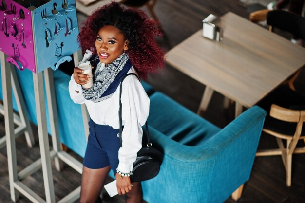 Attraktives afroamerikanisches lockiges Mädchen in weißer Bluse und blauen Shorts posierte im Café mit Latte und Handy zur Hand Ansicht von oben