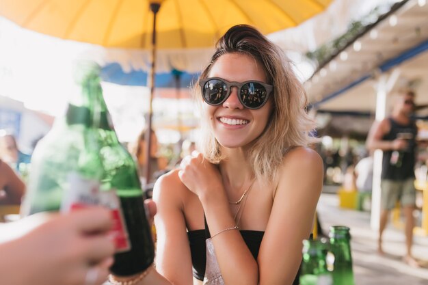 Attraktives blondes Mädchen, das Bier im Café trinkt und lacht. Aufgeregte junge Frau, die Zeit mit Freund im Restaurant im Freien am Sommertag verbringt.