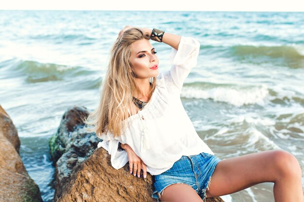 Attraktives blondes Mädchen mit langen Haaren sitzt auf Stein auf Seehintergrund.