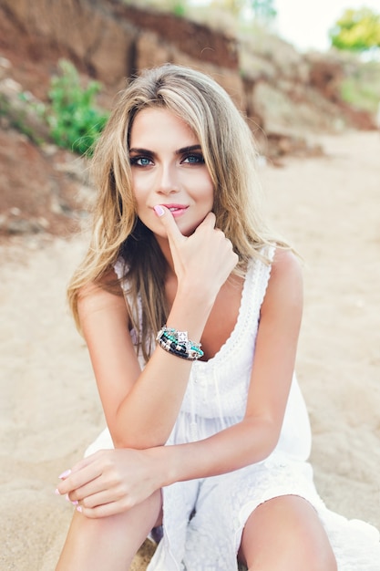 Attraktives blondes Mädchen mit langen Haaren und blauen Augen sitzt am Strand. Sie schaut in die Kamera.