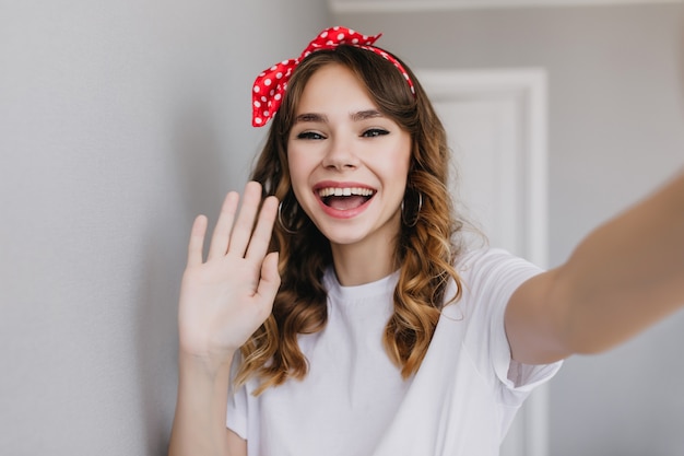 Attraktives brünettes Mädchen, das zu Hause mit fröhlichem Lächeln aufwirft. Bezauberndes europäisches weibliches Modell in Ohrringen, die Selfie machen.