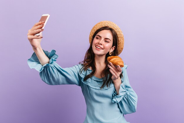 Attraktives dunkelhaariges Mädchen, das Croissant hält und Selfie auf lila Wand macht.