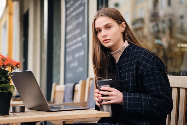 Attraktives ernstes Mädchen, das selbstbewusst in die Kamera schaut, während es sich mit Laptop im Café im Freien ausruht