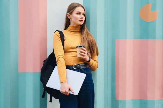 Attraktives ernsthaftes Studentenmädchen mit Laptop und Kaffee zum Mitnehmen, das über buntem Hintergrund im Freien wegschaut