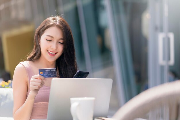 Attraktives erwachsenes asiatisches weibliches Glück, das online mit Laptophandgriff-Kreditkarte und Smartphone für Kaufprodukt mit Hintergrund des Geschäftsideenkonzeptes des Gebäudegartens im Freien einkauft