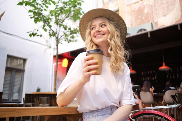 Attraktives, fröhliches, lässiges blondes Mädchen mit Hut, das freudig mit Kaffee zum Mitnehmen im Innenhof des Stadtcafés wegschaut