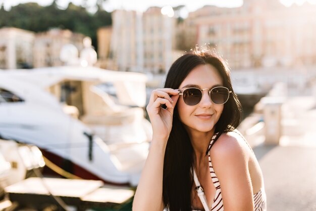 Attraktives fröhliches Mädchen mit Bronzehaut, das schwarze Sonnenbrille hält und draußen mit Booten ruht