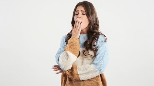 Attraktives gelangweiltes brünettes Mädchen im Pullover, das vor der Kamera über weißem Hintergrund gähnt