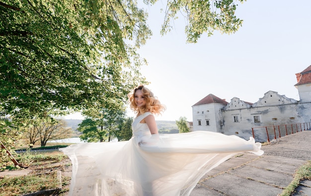 Attraktives glückliches blondes Mädchen gekleidet im weißen Kleid dreht sich um und lächelt am sonnigen Tag nahe altem Steinschloss