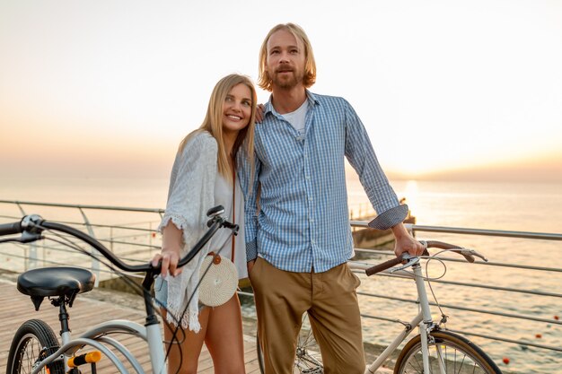 Attraktives glückliches Paar von Freunden, die im Sommer auf Fahrrädern reisen, Mann und Frau mit blondem Haar Boho-Hipster-Stil Mode, die Spaß zusammen haben, am Meer im Urlaub spazieren gehen