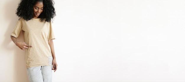 attraktives junges afrikanisches weibliches Modell mit stilvollem lockigem Haarschnitt, lässig gekleidet, fröhlich lächelnd, ihr T-Shirt mit Finger zeigend.