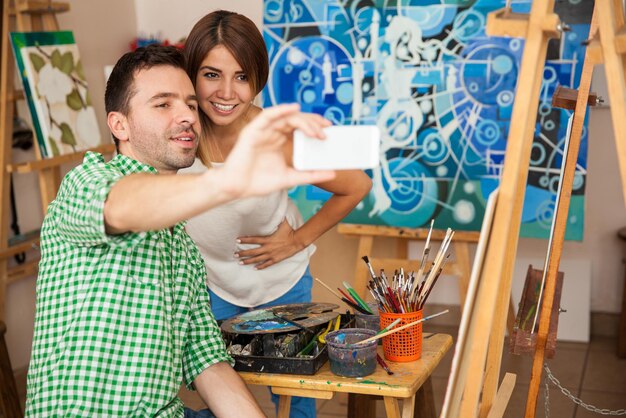 Attraktives junges hispanisches Paar, das ein Selfie mit einem Smartphone macht, während es bei einem Date an einem Kunstworkshop teilnimmt