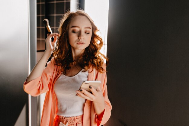Attraktives kaukasisches Mädchen im Pyjama, das mit Telefon zu Hause aufwirft. Innenfoto des kühlen weiblichen Modells mit welligem Haar, das Make-up-Pinsel hält.