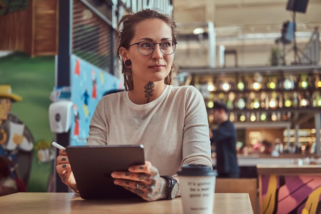 Kostenloses Foto attraktives kreatives mädchen mit tätowierungen an den händen sitzt im café und skizziert in ihrem digitalen notizblock.