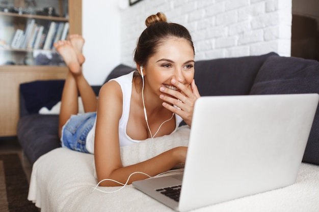 Attraktives lachendes junges Mädchen, das auf Sofa am Wohnzimmer mit Laptop liegt.