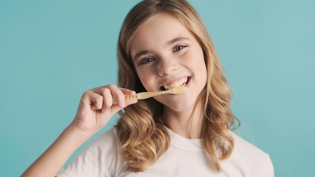 Attraktives lächelndes blondes Teenager-Mädchen, das ihre Zähne vor der Kamera putzt und glücklich über blauem Hintergrund aussieht