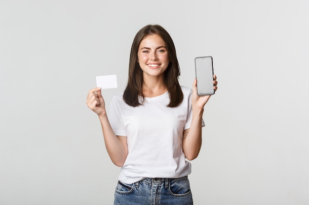Attraktives lächelndes Mädchen, das zufrieden schaut und Kreditkarte, Handybildschirm zeigt.