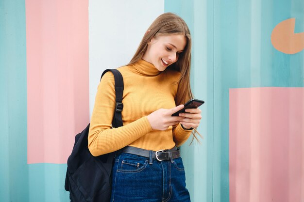 Attraktives lächelndes Studentenmädchen, das freudig Mobiltelefon über buntem Hintergrund im Freien verwendet