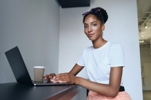 Attraktives lässiges afroamerikanisches Mädchen, das am Laptop mit Kaffee zum Mitnehmen arbeitet und selbstbewusst in die Kamera schaut in einem modernen Büro