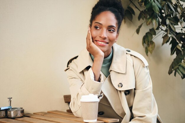 Attraktives lässiges afroamerikanisches Mädchen im stilvollen Trenchcoat, das glücklich mit Kaffee wegschaut, um im Café auf der Straße zu gehen