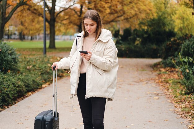 Attraktives lässiges Mädchen in Daunenjacke mit Koffer, das nachdenklich auf dem Handy im Freien aussieht