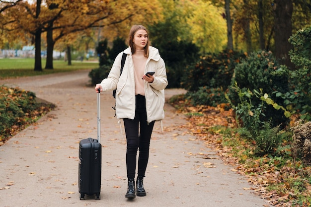 Attraktives lässiges Mädchen in Daunenjacke mit Koffer und Handy, das durch den Stadtpark geht