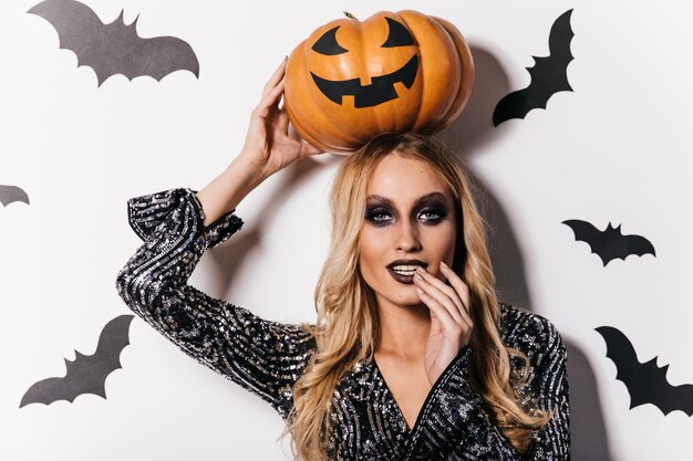 Attraktives langhaariges Mädchen, das auf weißer Wand mit Fledermäusen steht. Innenfoto des ansprechenden Vampirs mit Halloween-Kürbis.