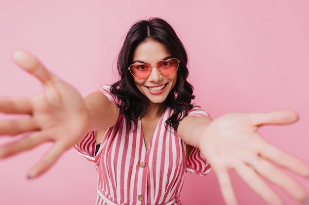 Attraktives lateinamerikanisches mädchen in der trendigen brille, die mit vergnügen und lachen aufwirft. innenaufnahme der faszinierenden frau in der gestreiften rosa kleidung.