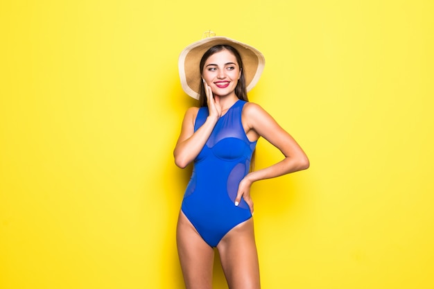 Attraktives Mädchen in einem weißen Bikini, Hut, Sonnenbrille, emotional geöffnetem Mund an einer gelben Wand mit einem perfekten Körper. Isoliert.